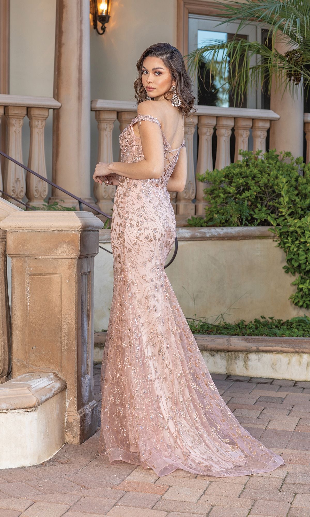 A woman in an off-the-shoulder long glitter prom dress stands on a paved path near a staircase and balustrade, looking over her shoulder.